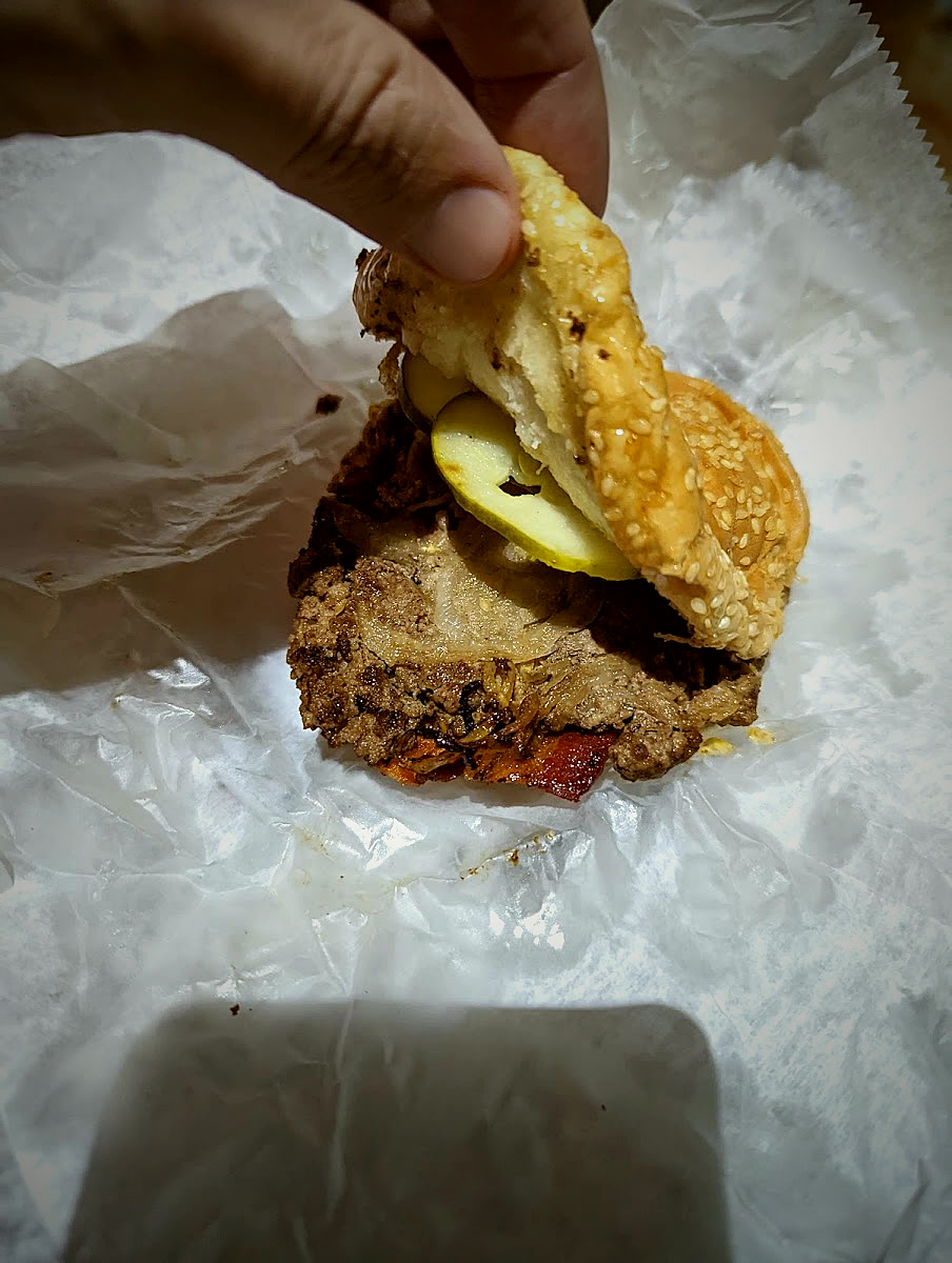 A hand lifting the top bun off a Geneva's burger to show the patty and pickles.
