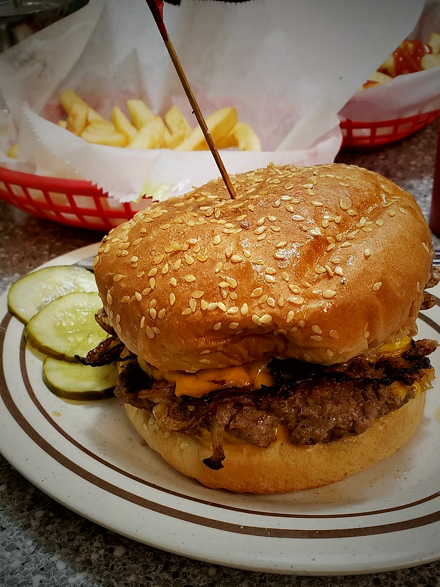 A Geneva's cheeseburger with pickles on a diner plate, a red basket of fries behind it.