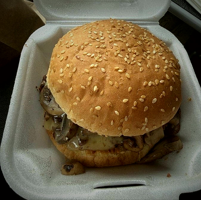 A mushroom and Swiss burger on a sesame bun in a takeout container.